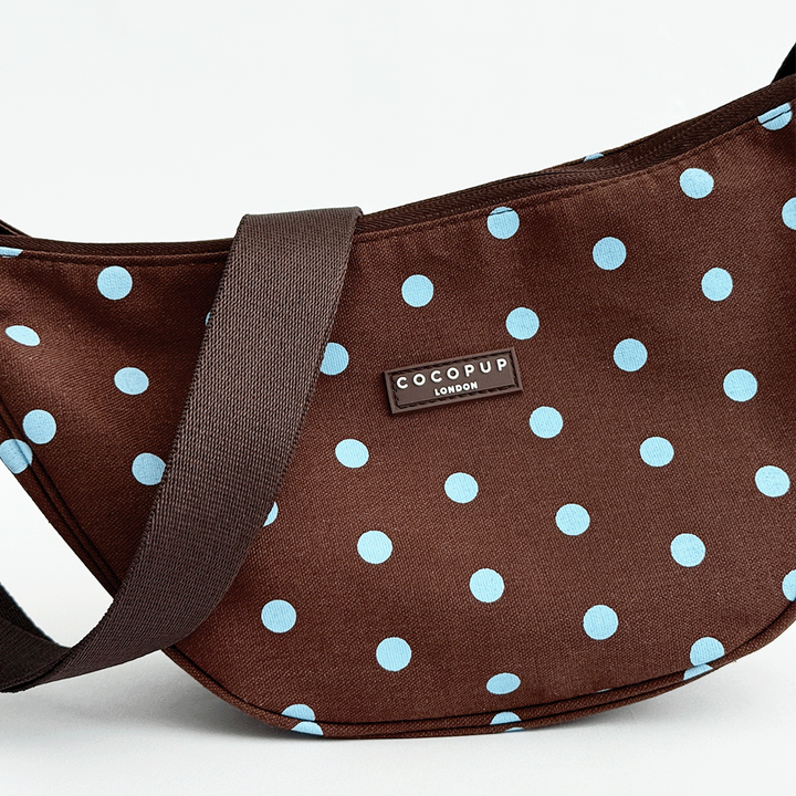 A brown fabric crossbody bag with a light blue polka dot pattern. Featuring Cocopup London logo badge and plain brown strap.