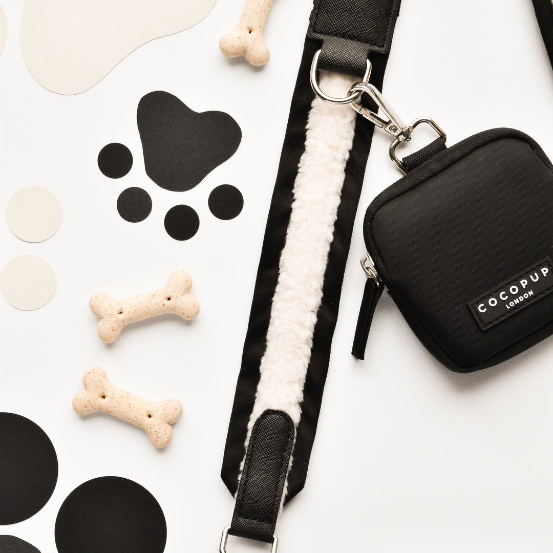 A flat lay image showcases a black dog leash with a white padded handle, a small black bag labeled "Cocopup London" featuring the "Bag Strap - Black Aviator," several bone-shaped dog treats, and paw print cutouts on a light background.