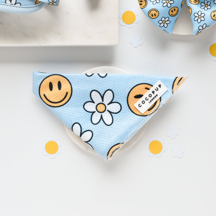 A light blue "Smiley Pawty Tent Bandana" pet bandana from Cocopup London, adorned with smiley faces and white daisies, is neatly folded on a white surface. Surrounded by scattered paper cut-outs of yellow-centered daisies, this playful accessory promises to add a cheerful flair to any pup.