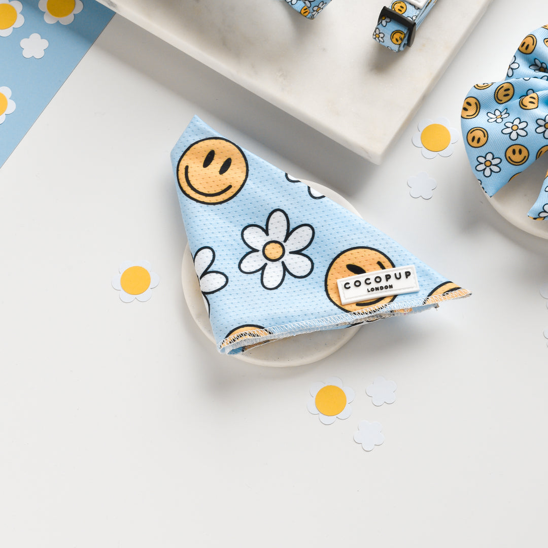 A light blue square cloth with a pattern of yellow smiley faces and white daisies is folded neatly on a marble surface. The playful Smiley Pawty Tent Bandana has a label on it that reads "Cocopup London." Small daisy paper cutouts are scattered around the scene, adding to the cheerful vibe.