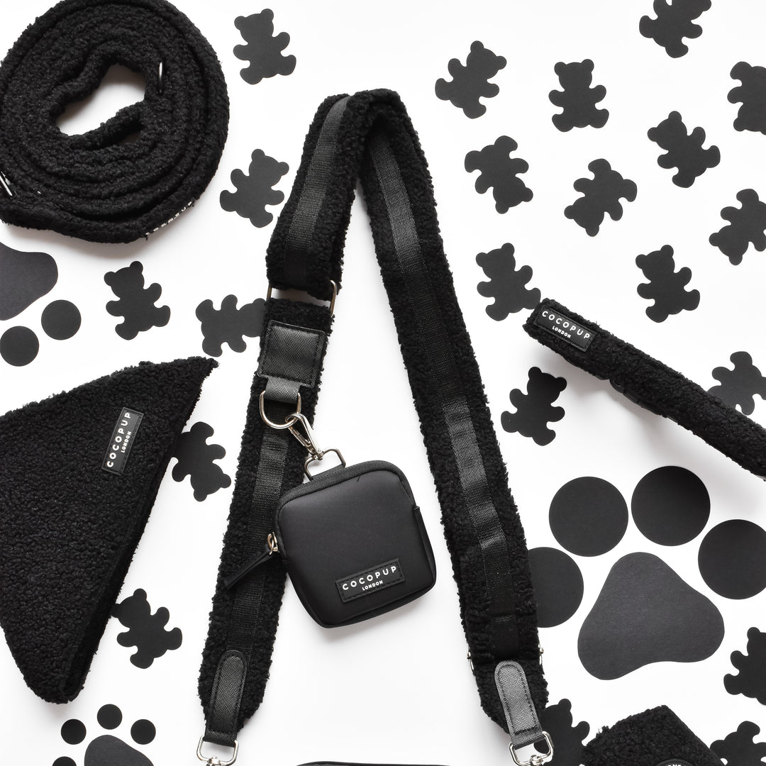 A neatly arranged collection of black dog accessories against a white background featuring black paw prints and dog bone shapes. Items include the Cocopup London Teddy Bag Strap - Baloo, leash with pouch, bandana, harness, and a fleece-lined collar with a fluffy design. Each item bears the "Cocopup London" label.