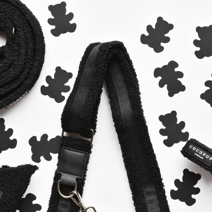 A black textured dog leash and harness are arranged on a white surface decorated with black teddy bear silhouettes. The harness features a metal clip and a label that reads "Cocopup London," complementing the fluffy design reminiscent of the Teddy Bag Strap - Baloo by Cocopup London.