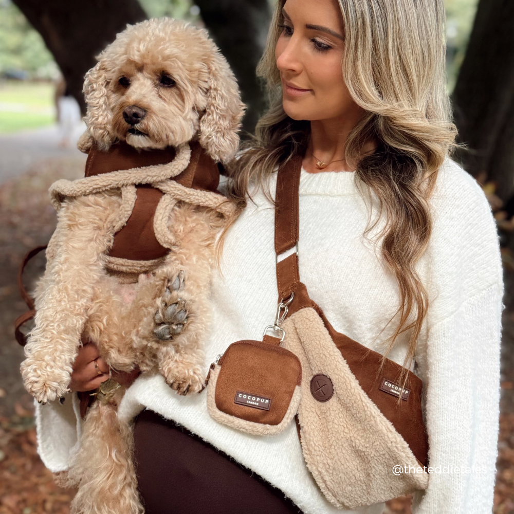 A woman in a white sweater carries a fluffy cream-colored dog wearing a brown harness while wearing a matching brown and cream crossbody dog bag with a small pouch. The brand name 'Cocopup' is visible on the bag's patch and strap label.