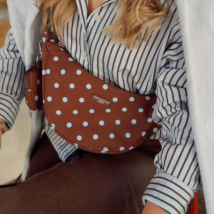 A brown crescent-shaped shoulder bag with light blue polka dots and a small brand plaque reading 'cocopup london', worn crossbody over a striped shirt and blue cardigan.