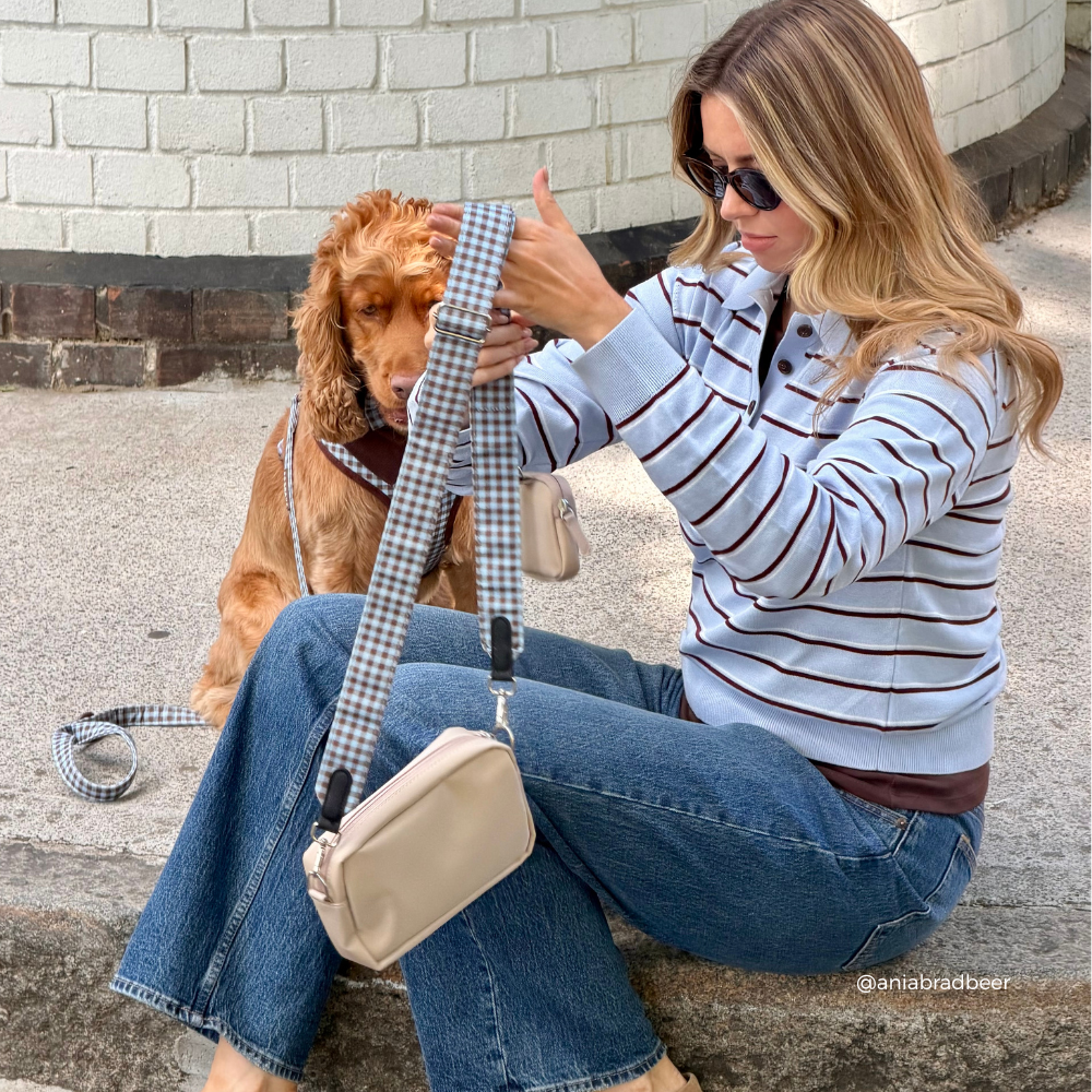 A woman sitting on concrete next to a brown dog outdoors, holding up a beige crossbody bag with a blue and brown gingham strap. She is wearing a light blue and brown striped long-sleeve top, blue jeans, and sunglasses.