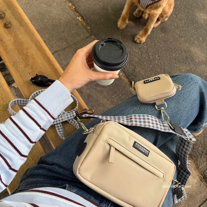 A person sitting outdoors on a bench wearing a beige crossbody bag with a gingham strap, a matching small beige pouch, and holding a takeaway coffee cup. A dog is visible in the background.
