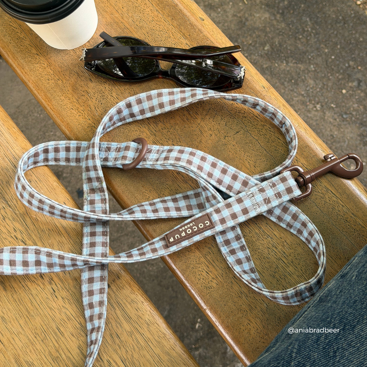 A brown and blue gingham check dog leash with a brown metal clasp, laid out on a wooden bench next to sunglasses, a coffee cup, and a person's jeans.