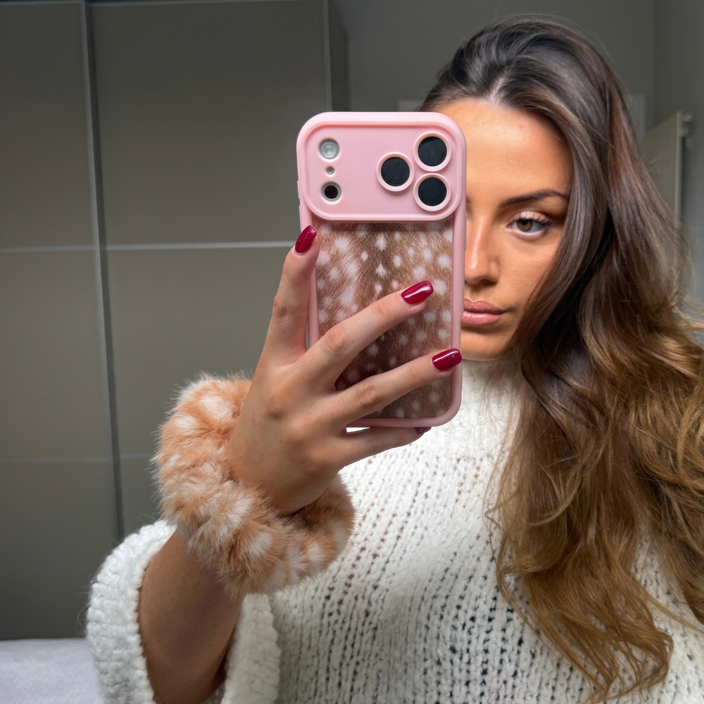 A woman takes a mirror selfie showing a pink phone case with a clear patterned back and a fluffy light brown wrist cuff. She is wearing a white knit sweater against a gray tiled bathroom background.