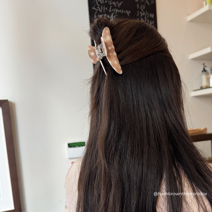 A person shown from behind with long dark brown hair, wearing a large fawn claw hair clip in a salon-like indoor setting.