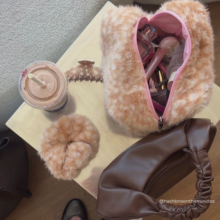 A styled flat-lay of a fluffy faux-fur cosmetic pouch in a warm beige and brown pattern, shown alongside a pink phone case, hair accessories, a perfume bottle, and an iced coffee on a carpeted floor.
