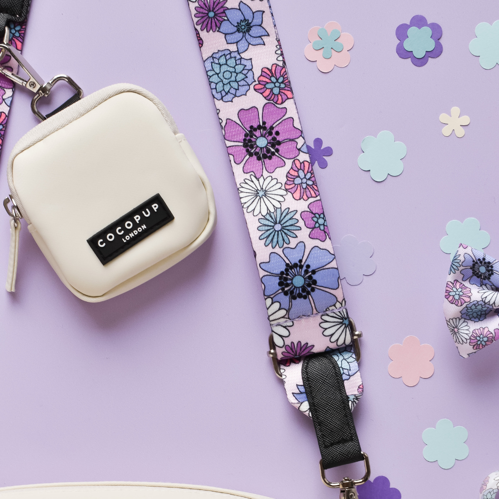 A small, square cream-colored dog walking bag featuring a black "Cocopup London" label is attached to the "Bag Strap - Pastel Flowers" from Cocopup London. The strap is adorned with pastel flowers in shades of purple, blue, and pink against a lavender background decorated with additional flower cutouts, enhancing the floral theme.